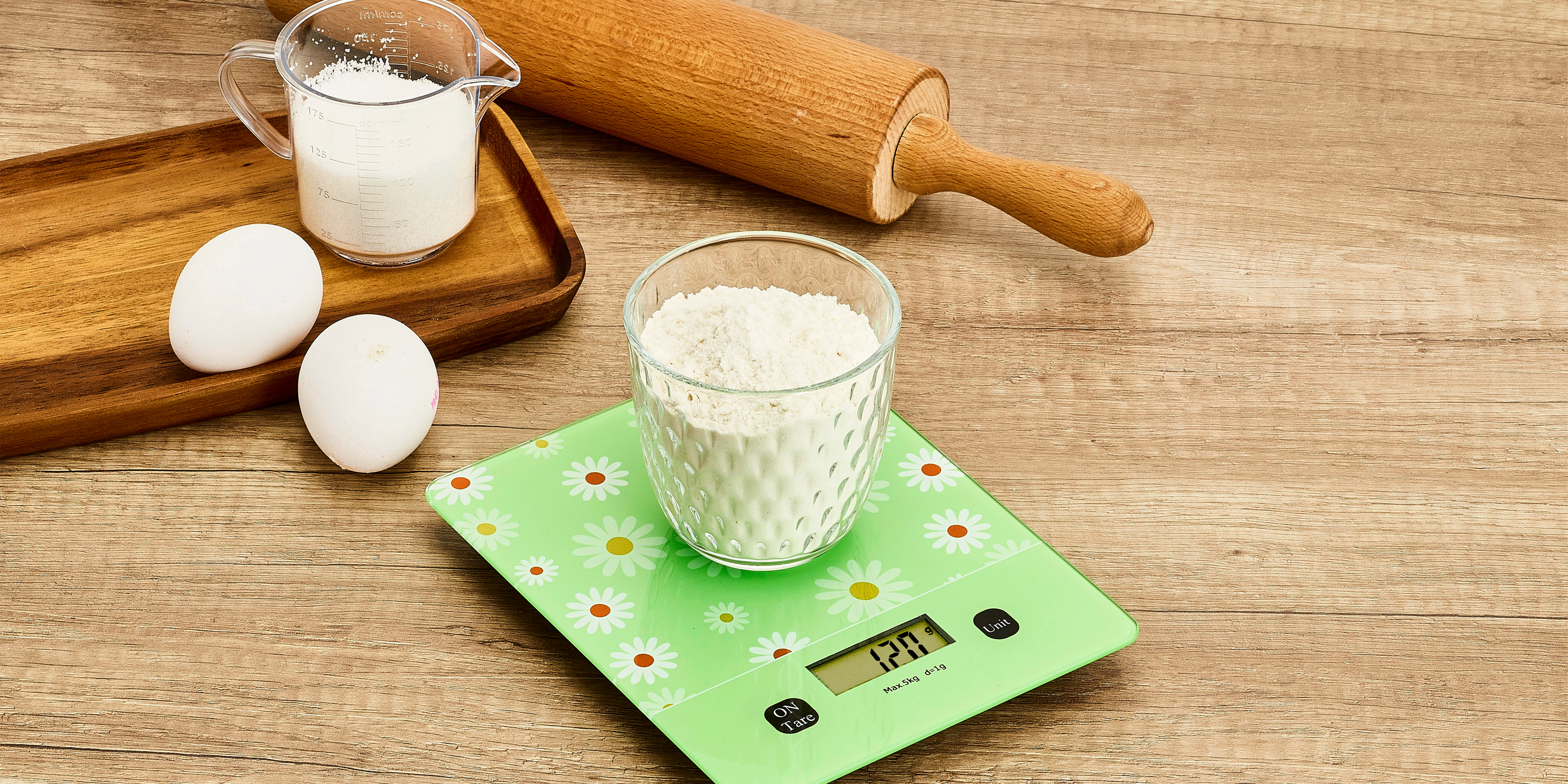Eine grüne Küchenwaage mit Blumendesign zeigt eine Schale mit Mehl, daneben zwei Eier, ein Messbecher mit Milch und ein Nudelholz auf einer Holzarbeitsfläche.