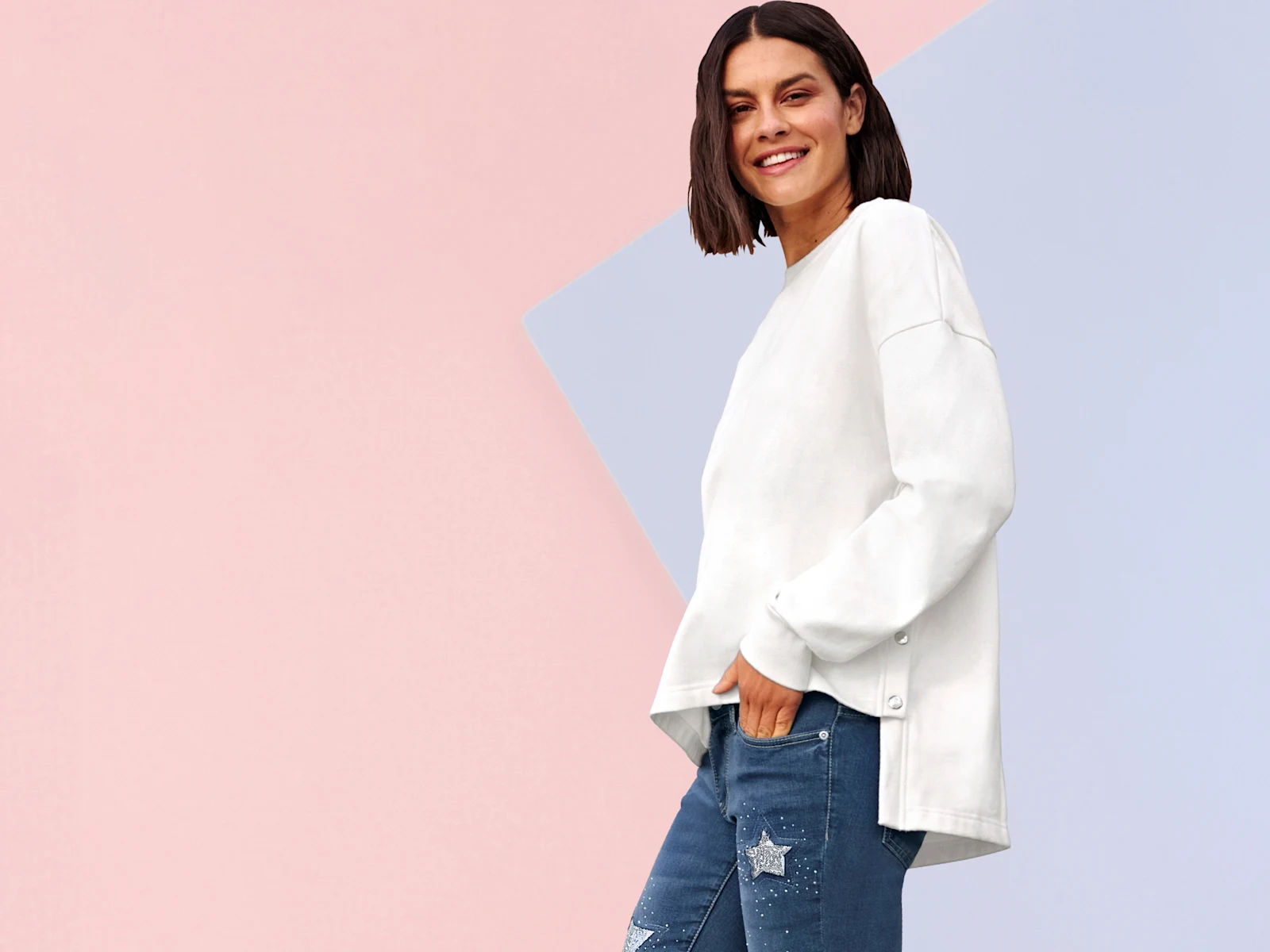 Model má na sebe biely voľný sveter so zapínaním na gombíky na boku a modré rifle s hviezdovými aplikáciami.