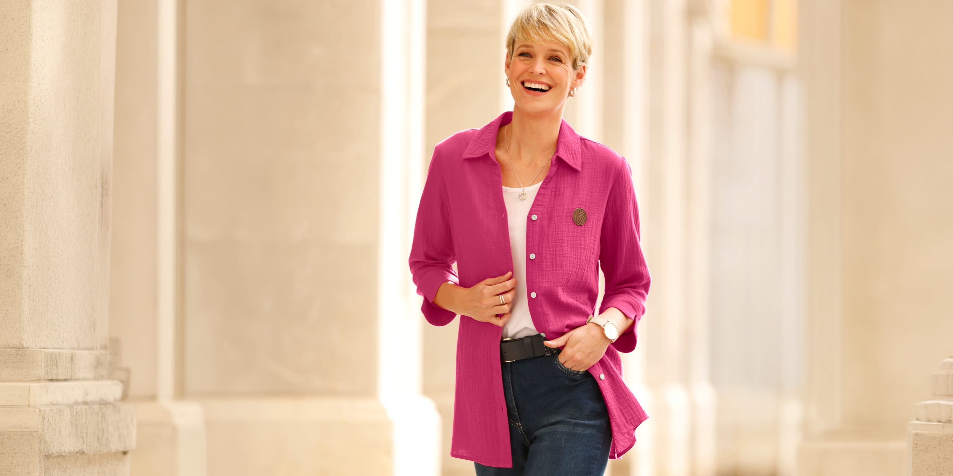 Model má na sebe ružovú dlhú blúzu cez biele tričko a modré džínsy.