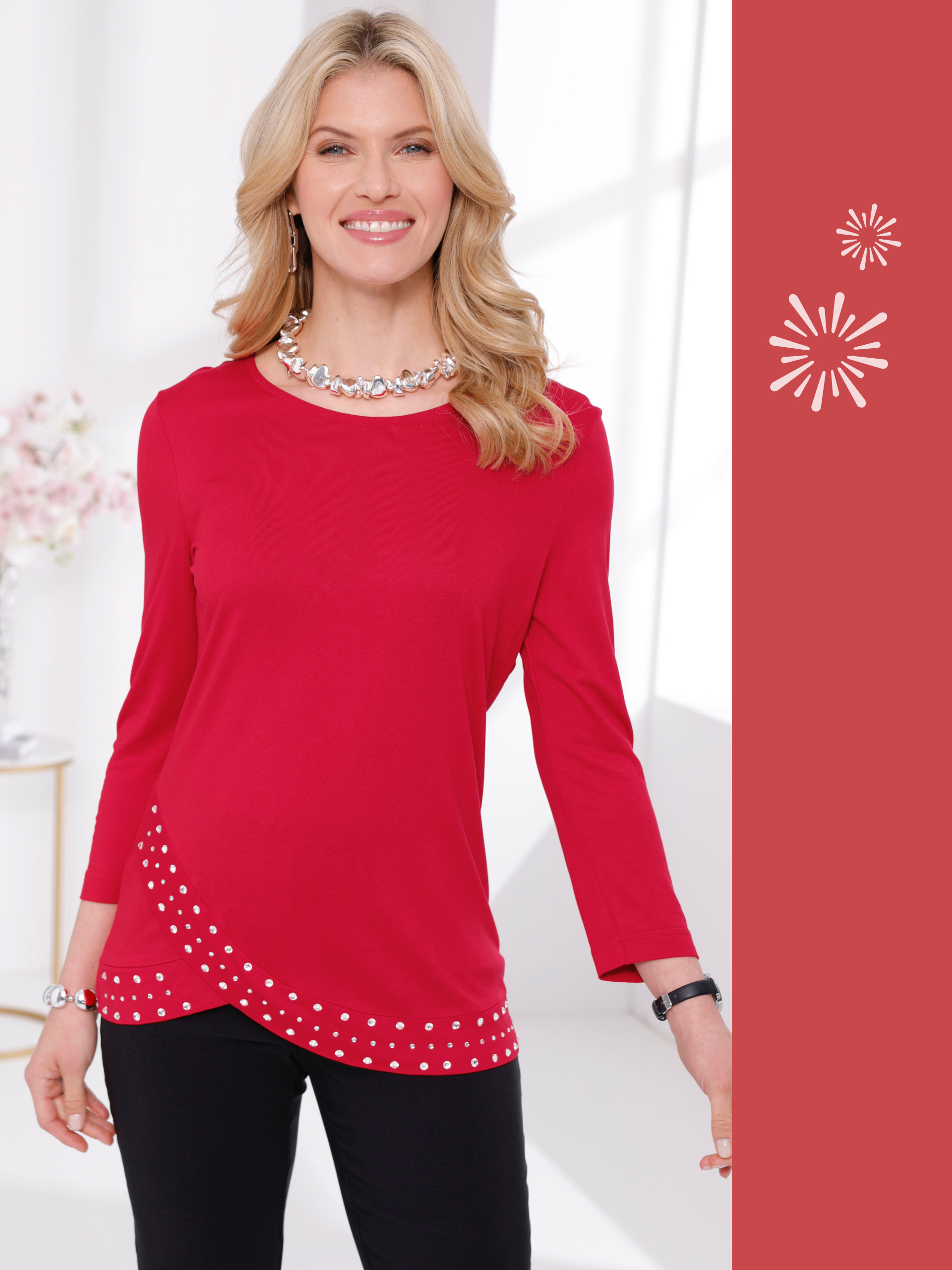 En modell med blont lockigt hår bär en röd blus med strassstenar, ett iögonfallande halsband och örhängen. Hon ler.