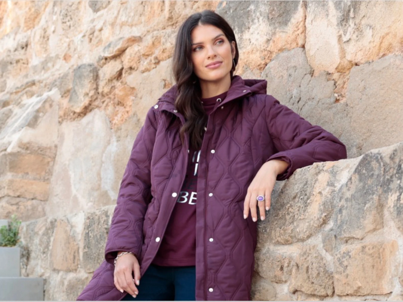 Modelka má oblečený bordový prešívaný kabát pred stenou z prírodného kameňa.