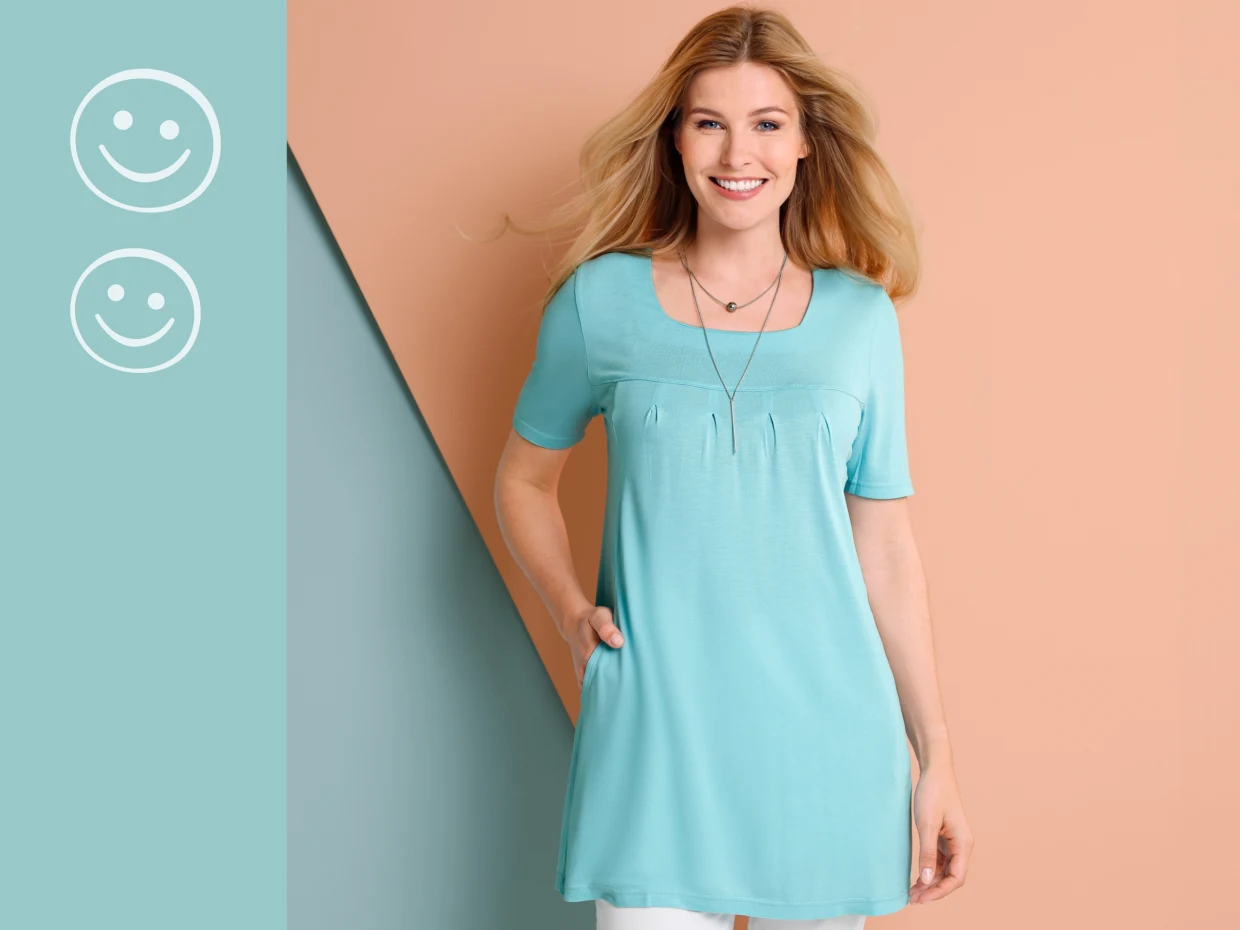 Damen-Tunika in Türkis mit kurzen Ärmeln, Rundhalsausschnitt und Falten-Detail, kombiniert mit weißer Hose vor zweifarbigem Hintergrund.