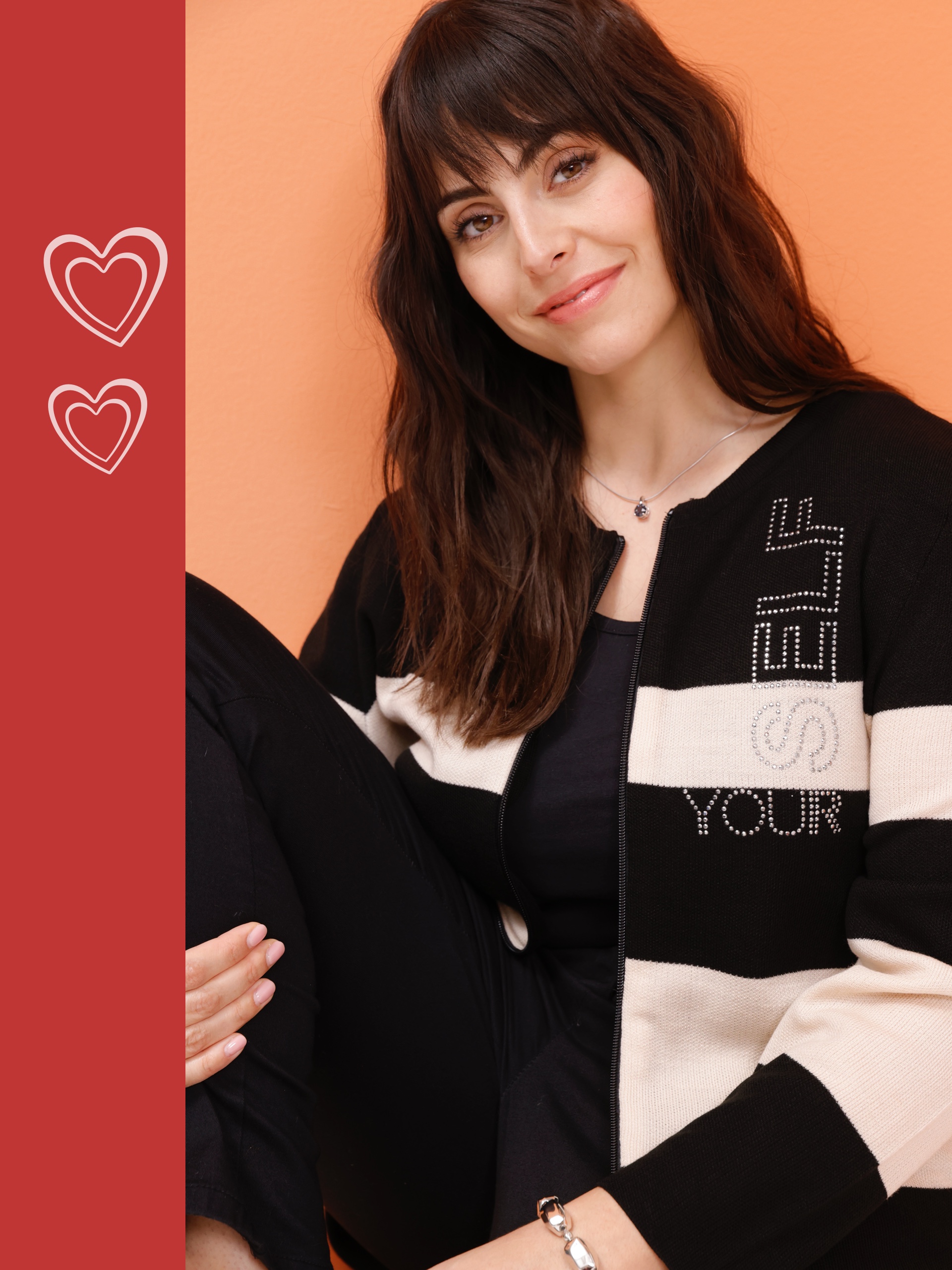 Ein Model sitzt in einem gestreiften, schwarz-weißen Cardigan und schwarzer Kleidung, vor einem orangefarbenen Hintergrund.