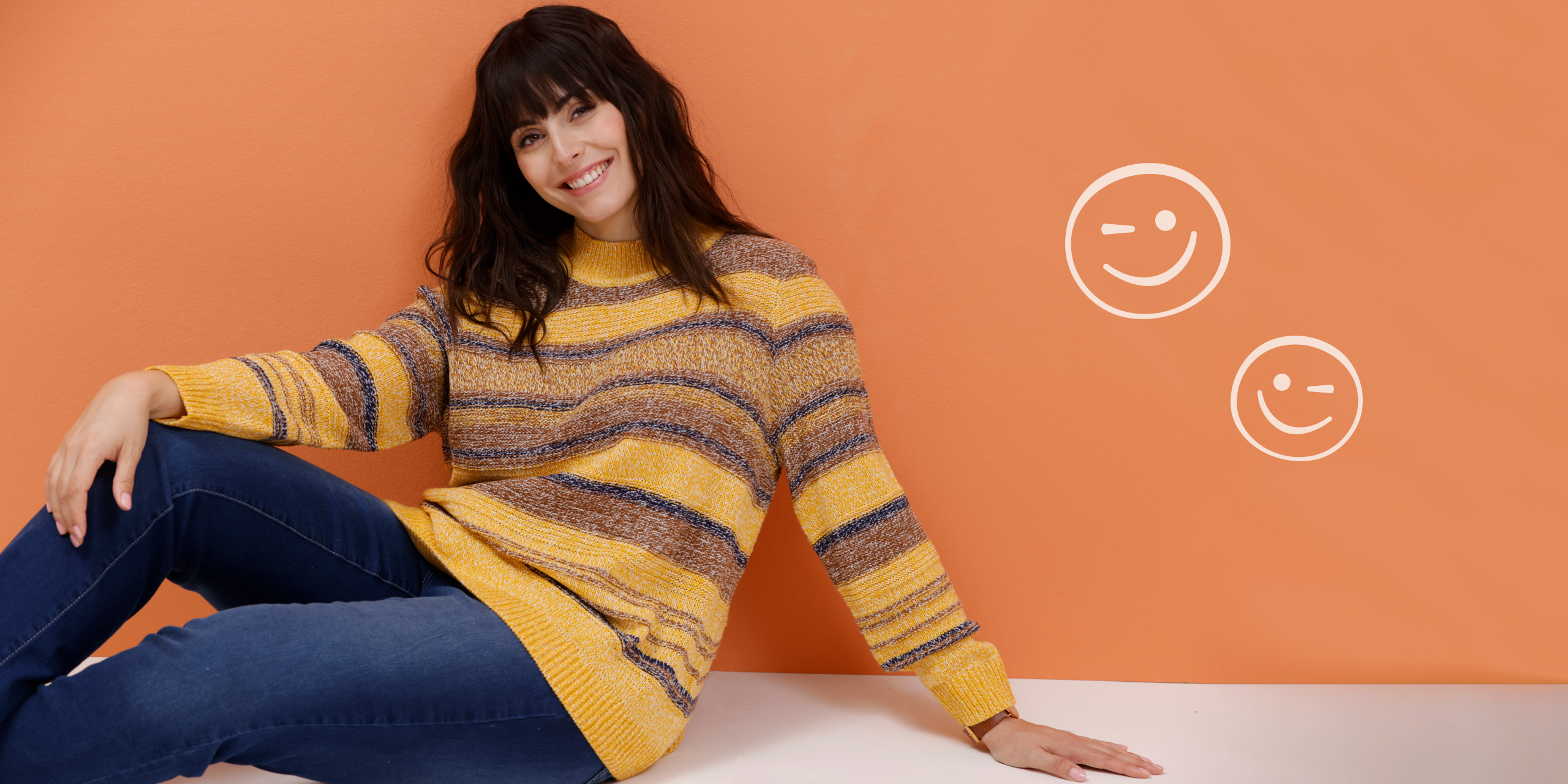 Ein Model sitzt vor einem orangefarbenen Hintergrund und präsentiert einen gestreiften Pullover in Gelb- und Brauntönen und eine blaue Jeans.