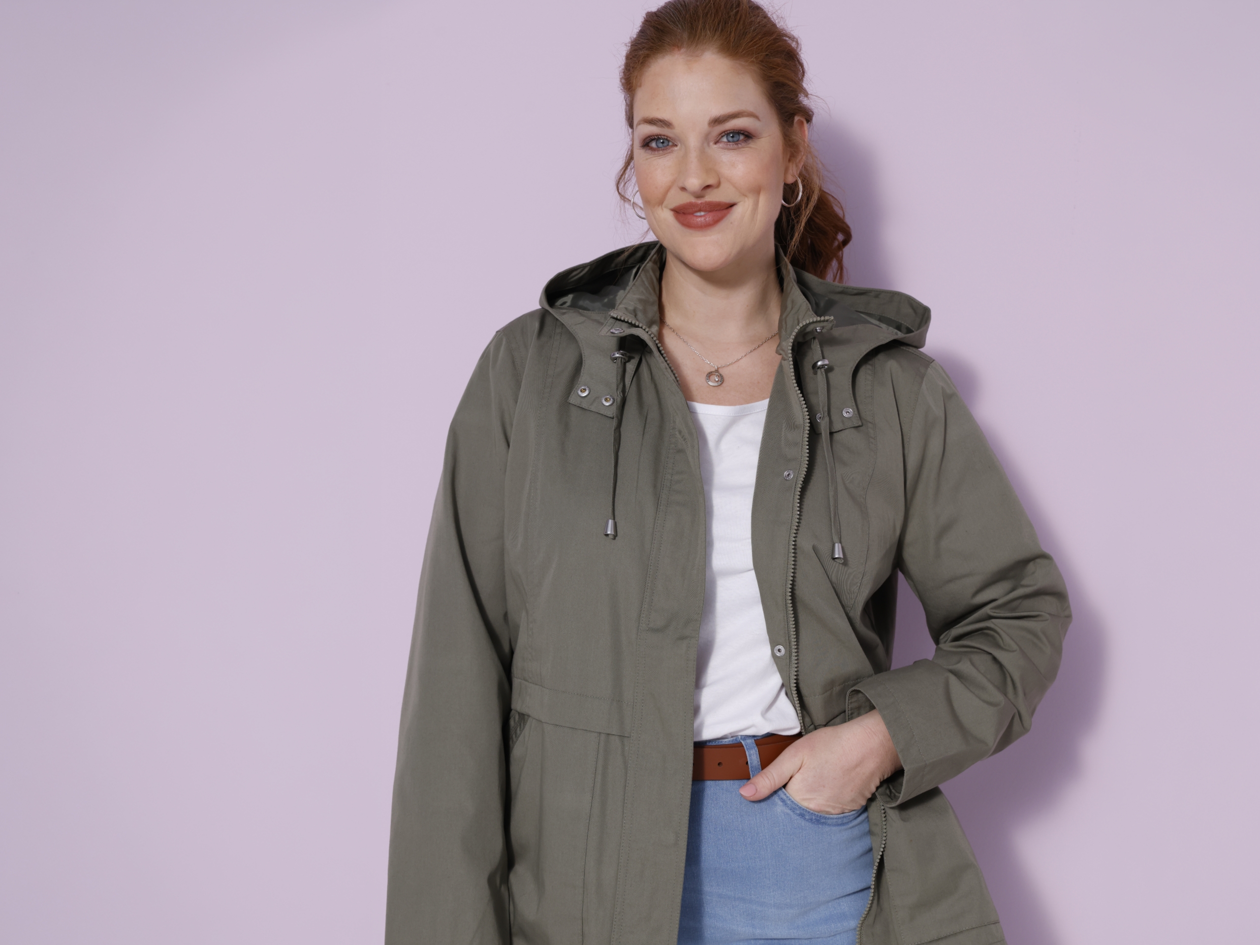 Een model draagt een olijfgroene capuchonjas over een wit T-shirt en een lichtblauwe spijkerbroek en staat voor een lichte achtergrond.