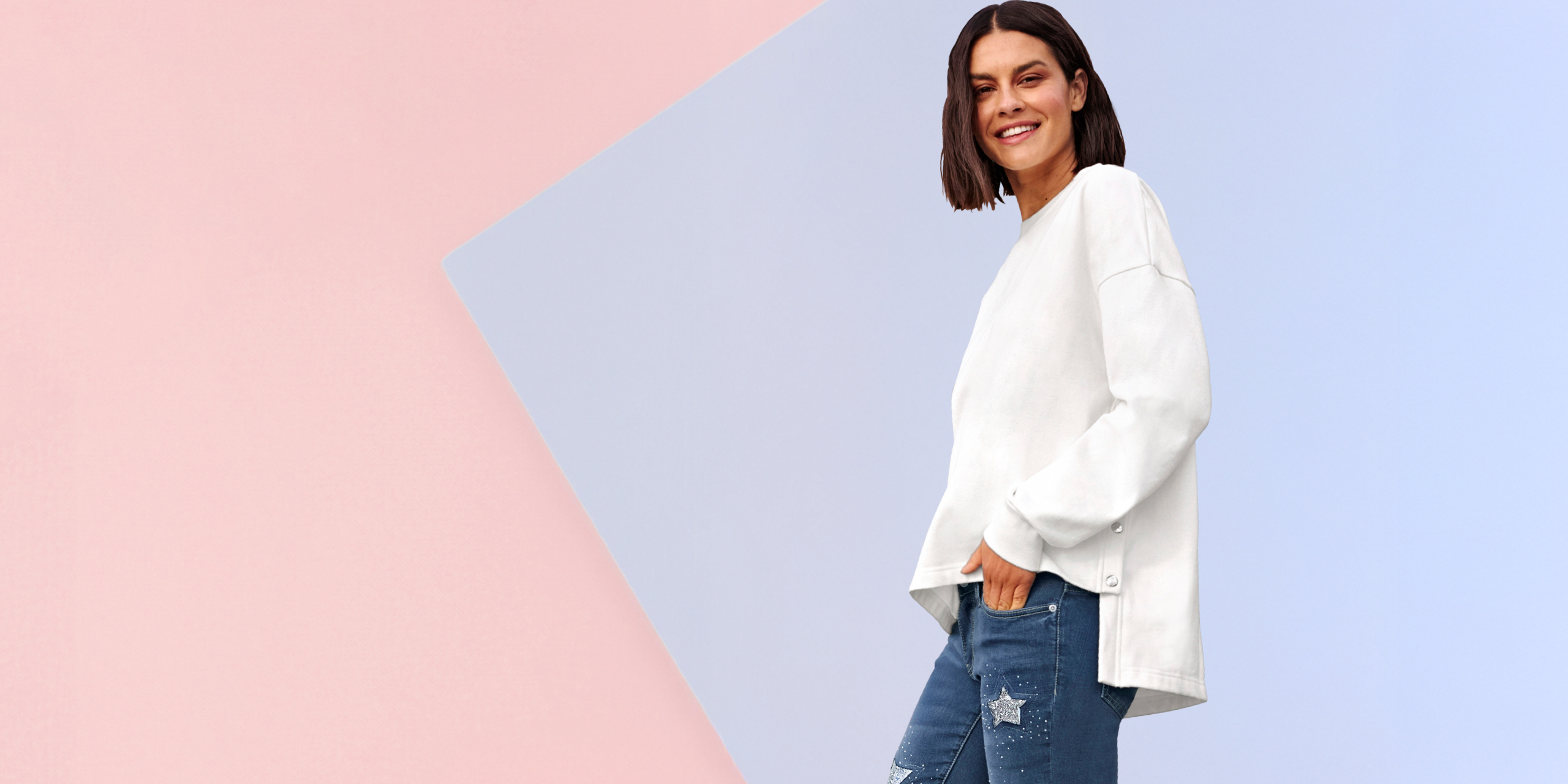 Model má na sebe biely voľný sveter so zapínaním na gombíky na boku a modré rifle s hviezdovými aplikáciami.