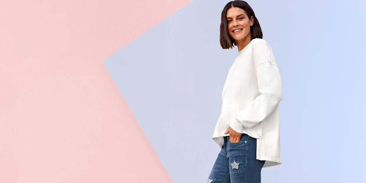 Model má na sebe biely voľný sveter so zapínaním na gombíky na boku a modré rifle s hviezdovými aplikáciami.