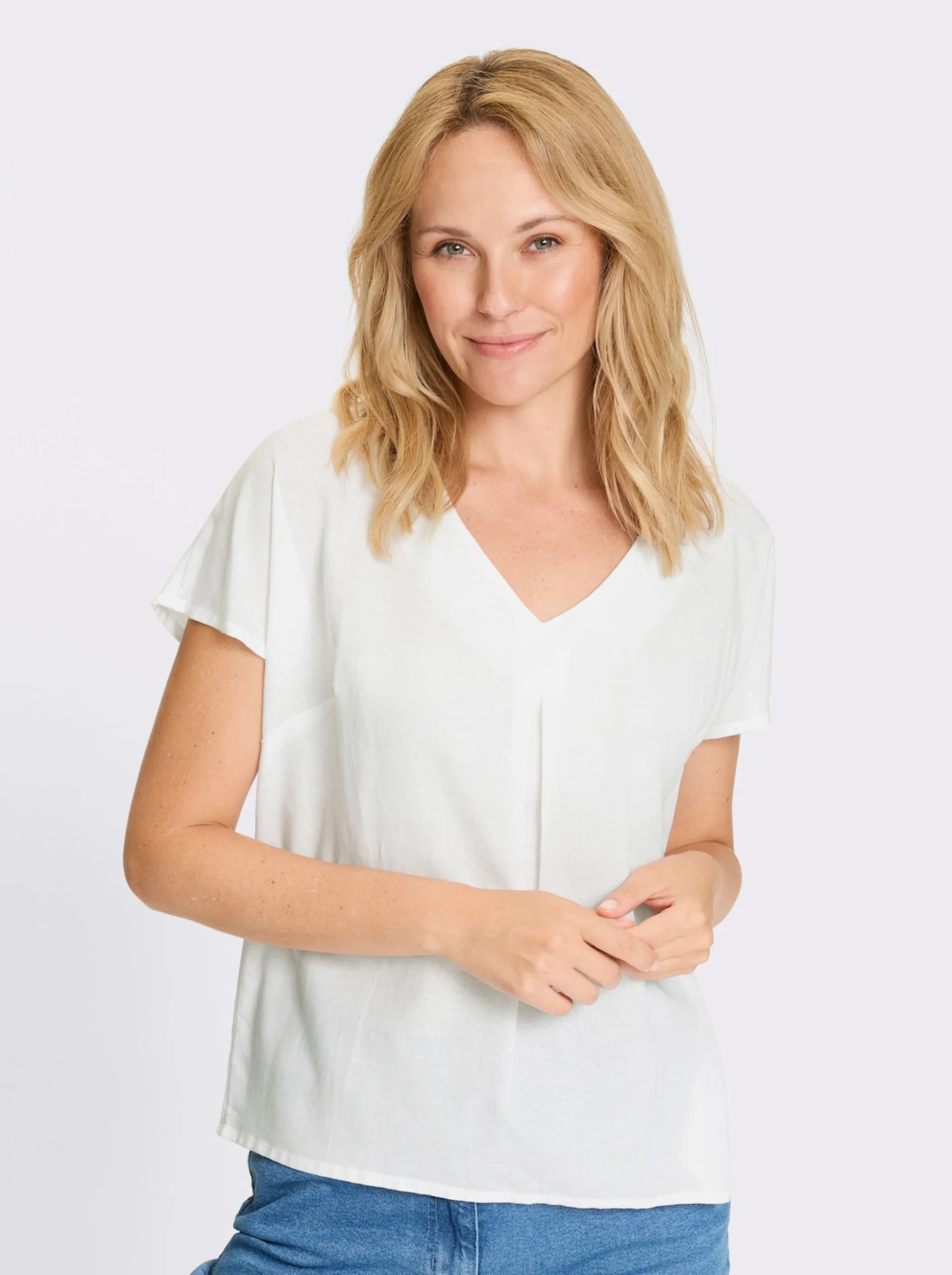 Frau trägt eine weiße Schlupfbluse im Viskose-Leinen-Mix. Mit aufspringender Falte unterhalb des V-Ausschnitts. Überschnittene Schultern und formgebende Brustabnäher. Frau trägt eine weiße Schlupfbluse im Viskose-Leinen-Mix. Mit aufspringender Falte unterhalb des V-Ausschnitts. Überschnittene Schultern und formgebende Brustabnäher.