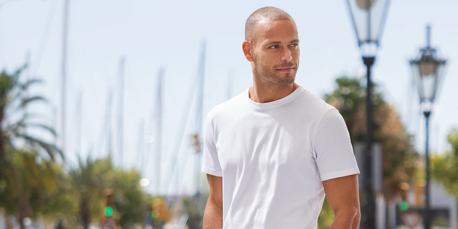 Een model met kort haar draagt een wit T-shirt en staat buiten voor een wazige achtergrond met bomen en lampen.