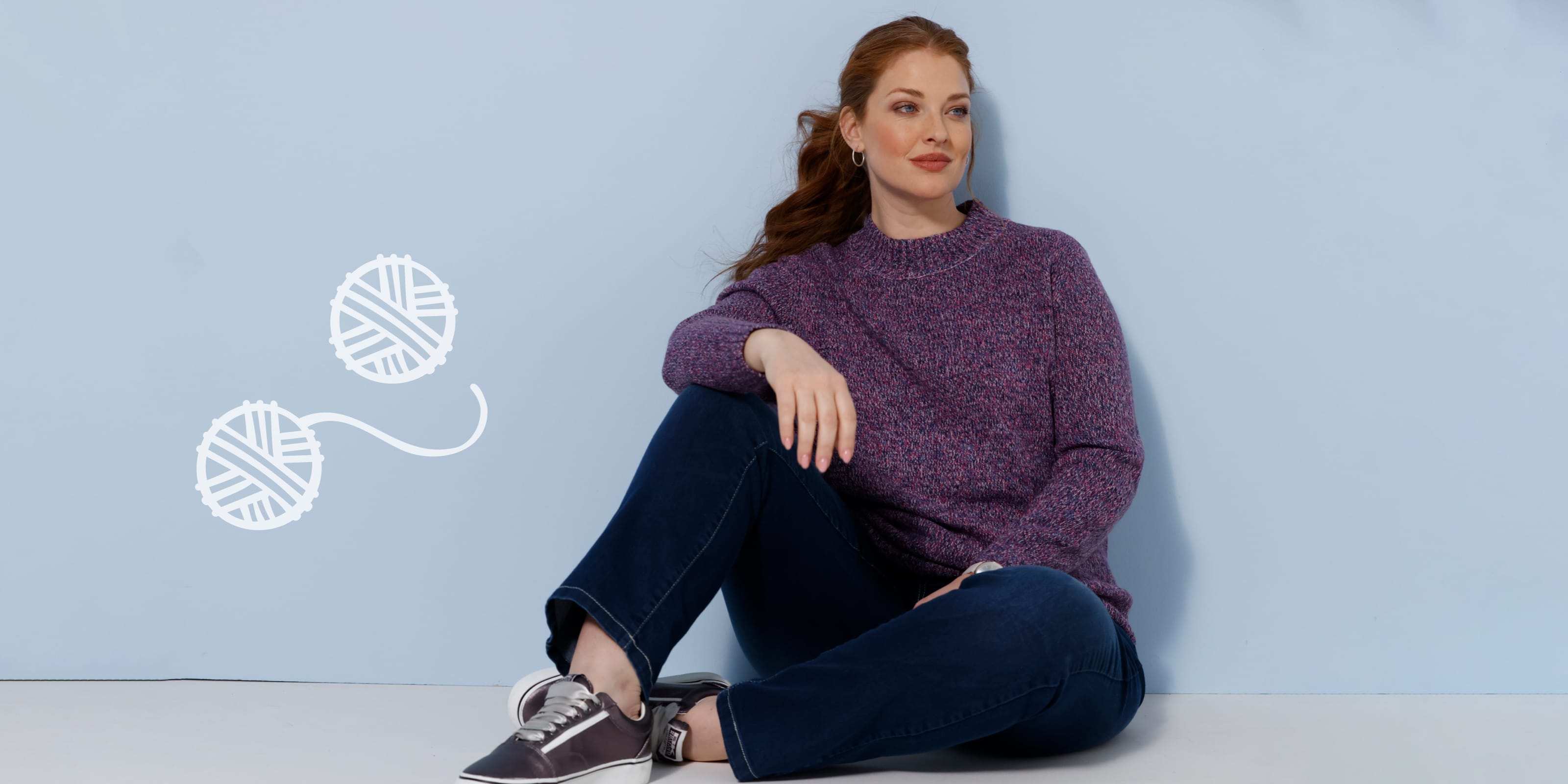 Ein Model sitzt in lila Strickpullover, Jeans und Turnschuhen vor hellblauem Hintergrund.