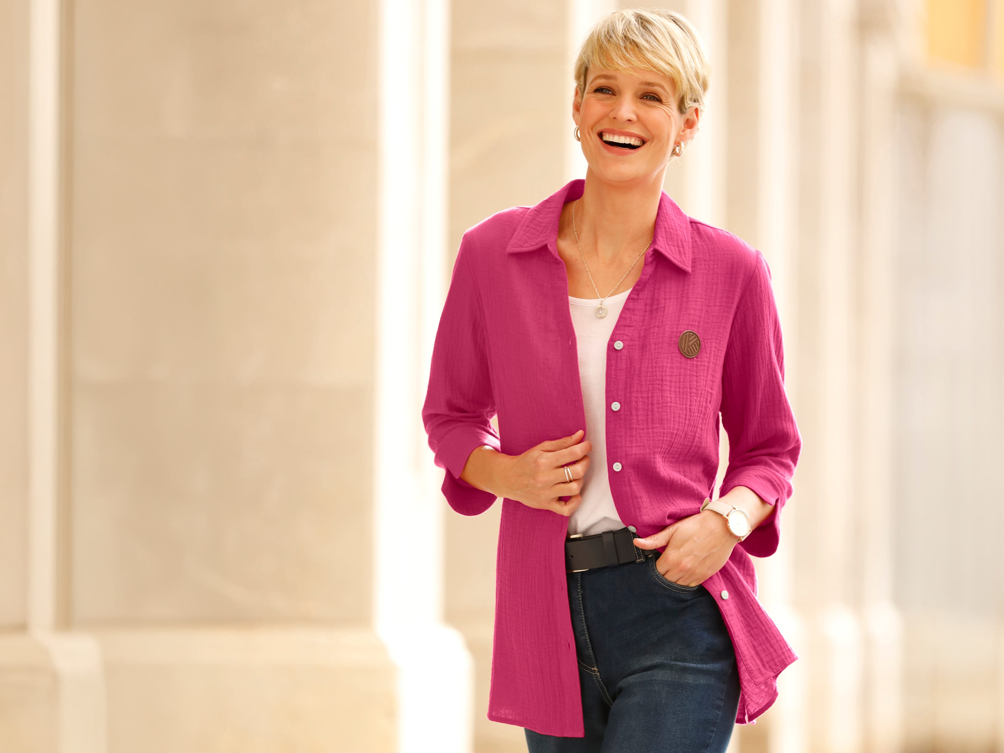 Model má na sebe ružovú dlhú blúzu cez biele tričko a modré džínsy.