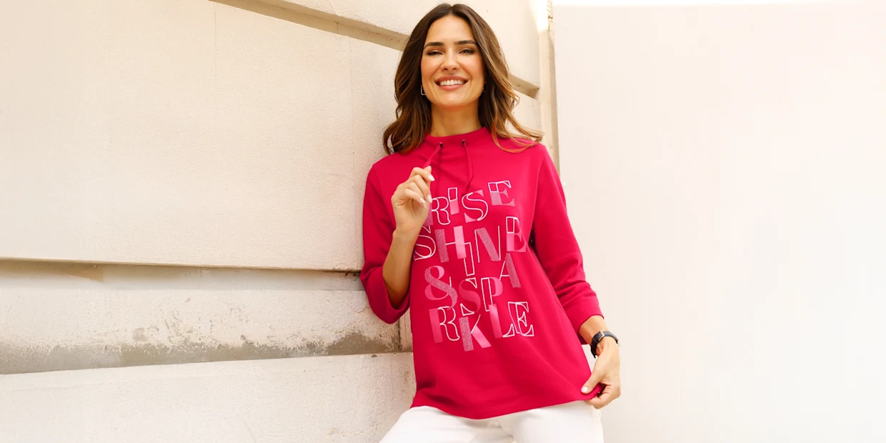 Modelka prezentuje ružovú mikinu s kapucňou s nápisom 'Rise Shine & Sparkle' a má na sebe biele nohavice.