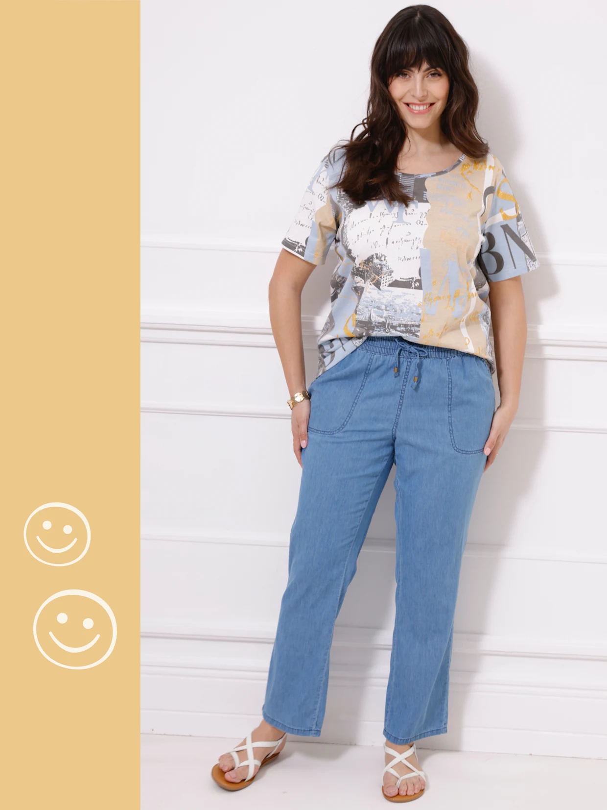 Ein Model trägt ein gemustertes T-Shirt und hellblaue Jeans, während es vor einer weißen Wand steht.