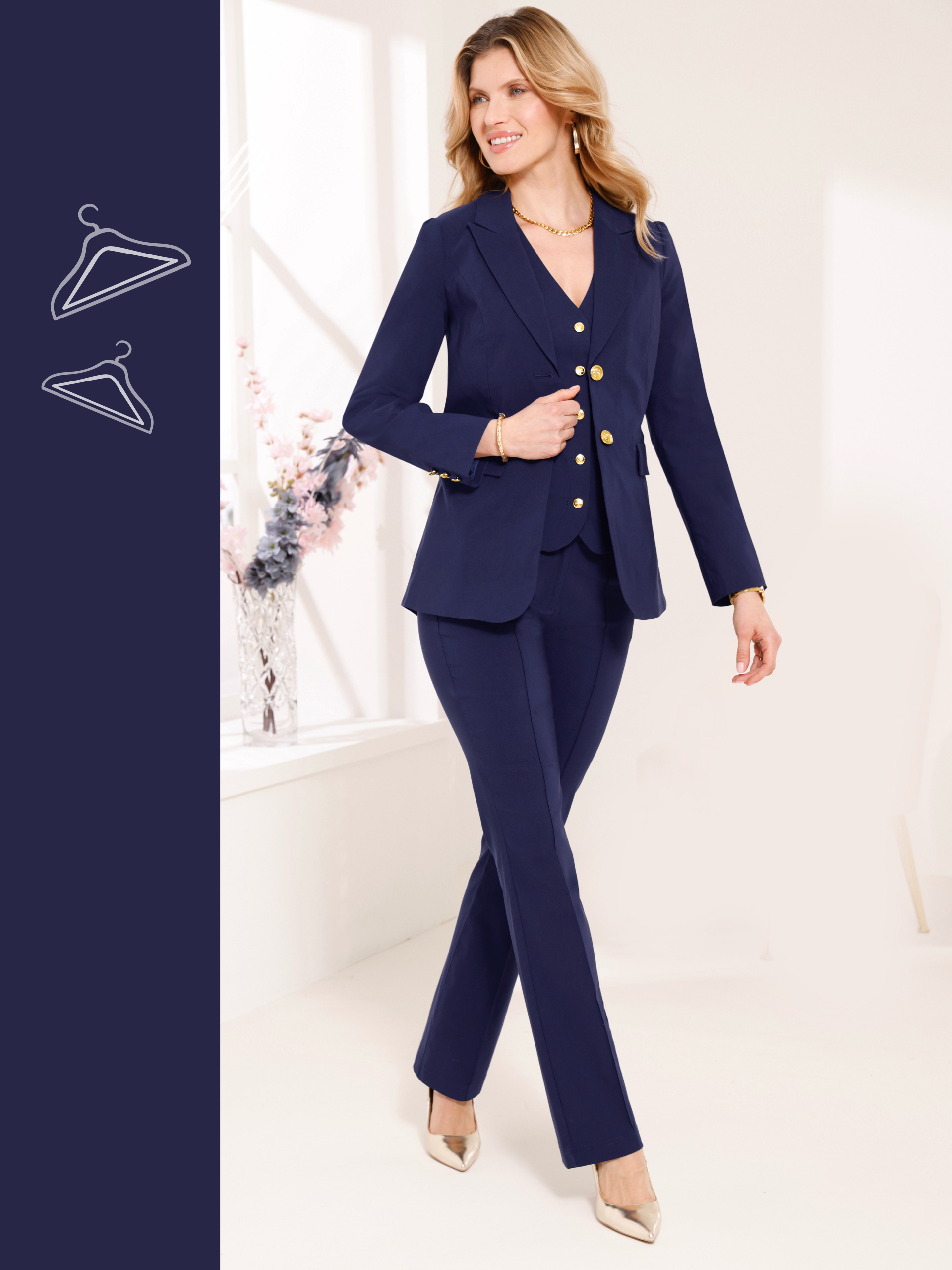Ein Model trägt einen dunkelblauen Hosenanzug mit goldfarbenen Knöpfen und goldenen Schuhen, steht in hellem Raum.