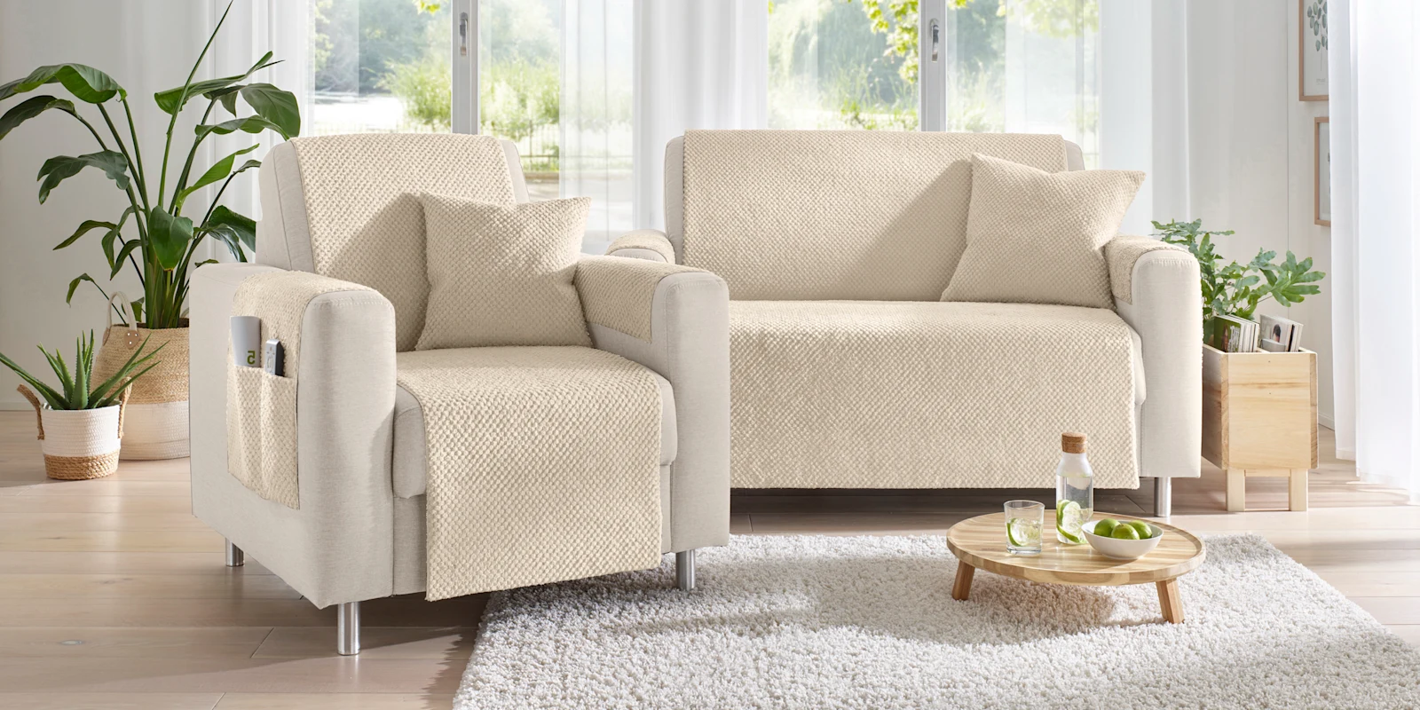 Ein hellbeiges Sofa und ein Sessel mit Überwürfen und Kissen stehen in einem hellen Wohnzimmer mit Pflanzen und Fensterfront.