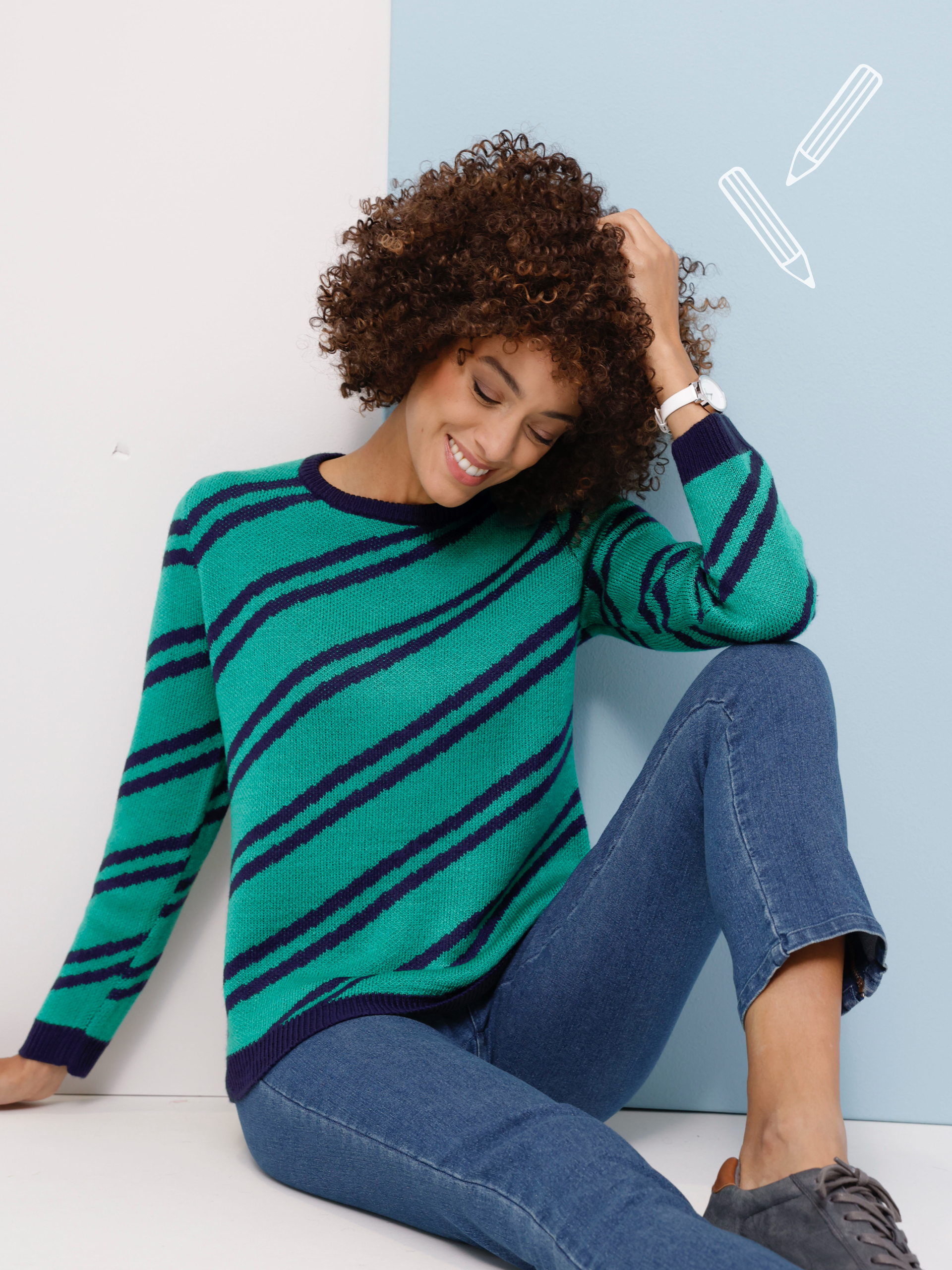 Model sedí a má na sebe zelený sveter so šikmými tmavomodrými pruhmi, modré džínsy a sivé topánky.