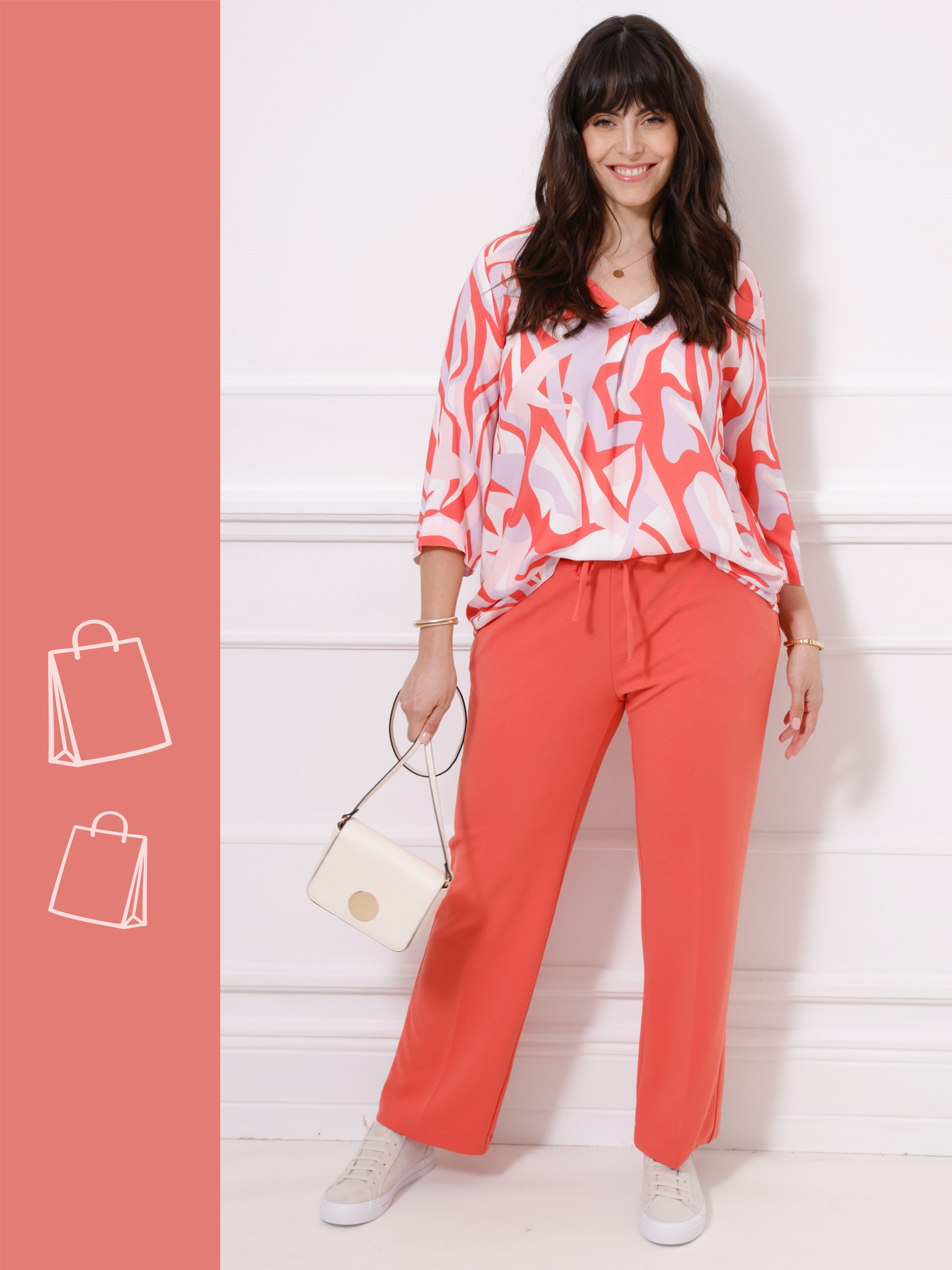 Frau trägt ein Outfit in Koralltönen bestehend aus gemusterter Bluse und weit geschnittenen Hosen, kombiniert mit weißer Umhängetasche und weißen Sneakers vor hellem Hintergrund.