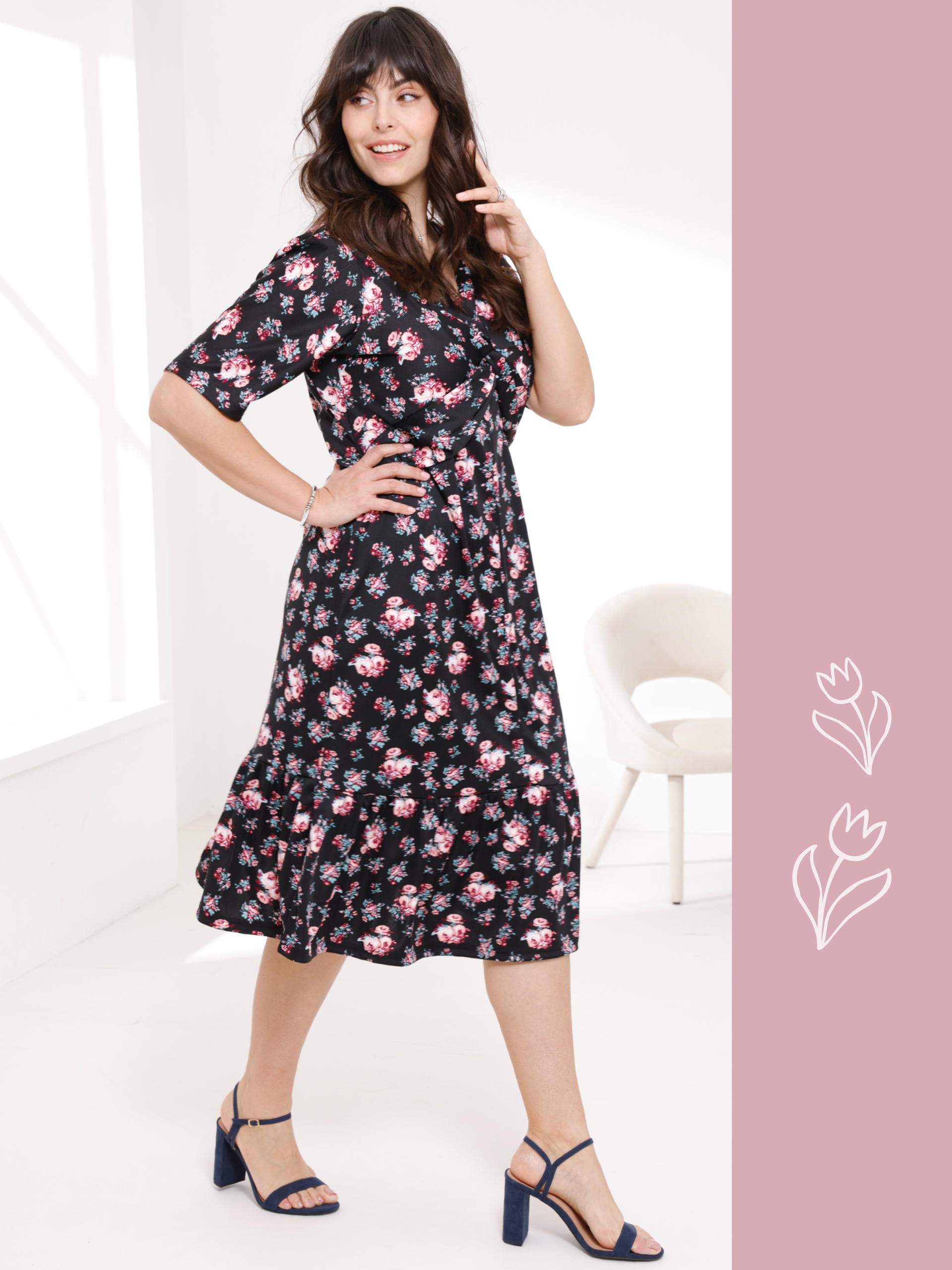 Ein Model trägt ein schwarzes, blumengemustertes Kleid und steht in einer Pose vor einem hellen Hintergrund.