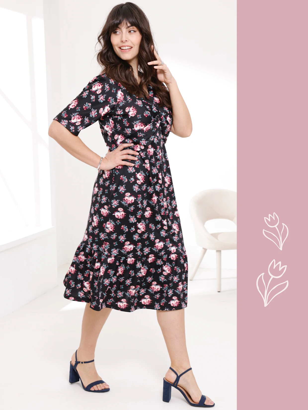 Ein Model trägt ein schwarzes, blumengemustertes Kleid und steht in einer Pose vor einem hellen Hintergrund.