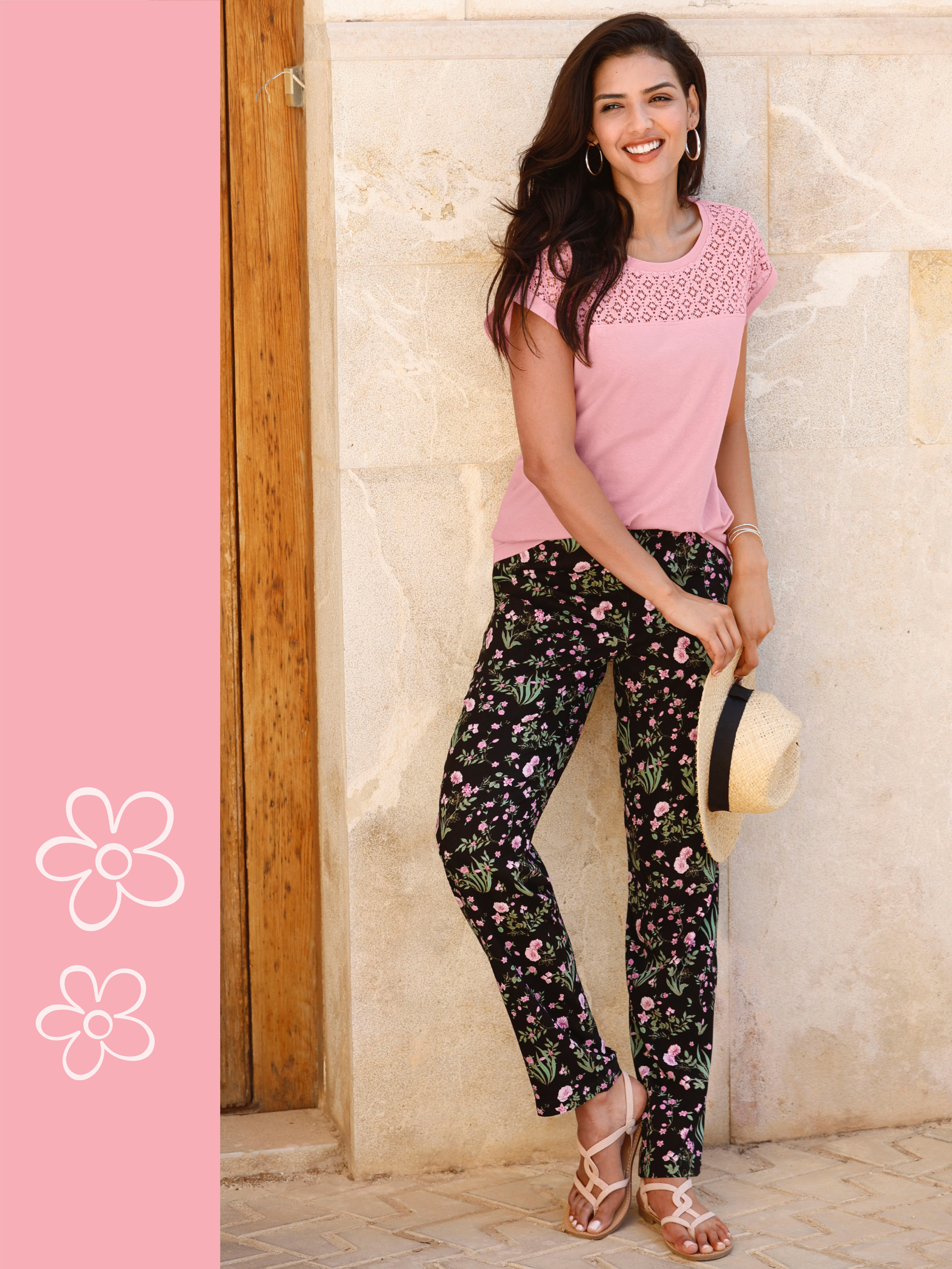 Frau trägt ein rosa T-Shirt mit Spitzenmuster und eine schwarze Hose mit floralem Print, kombiniert mit Sandalen und einem Strohhut, lehnt an einer Steinwand.