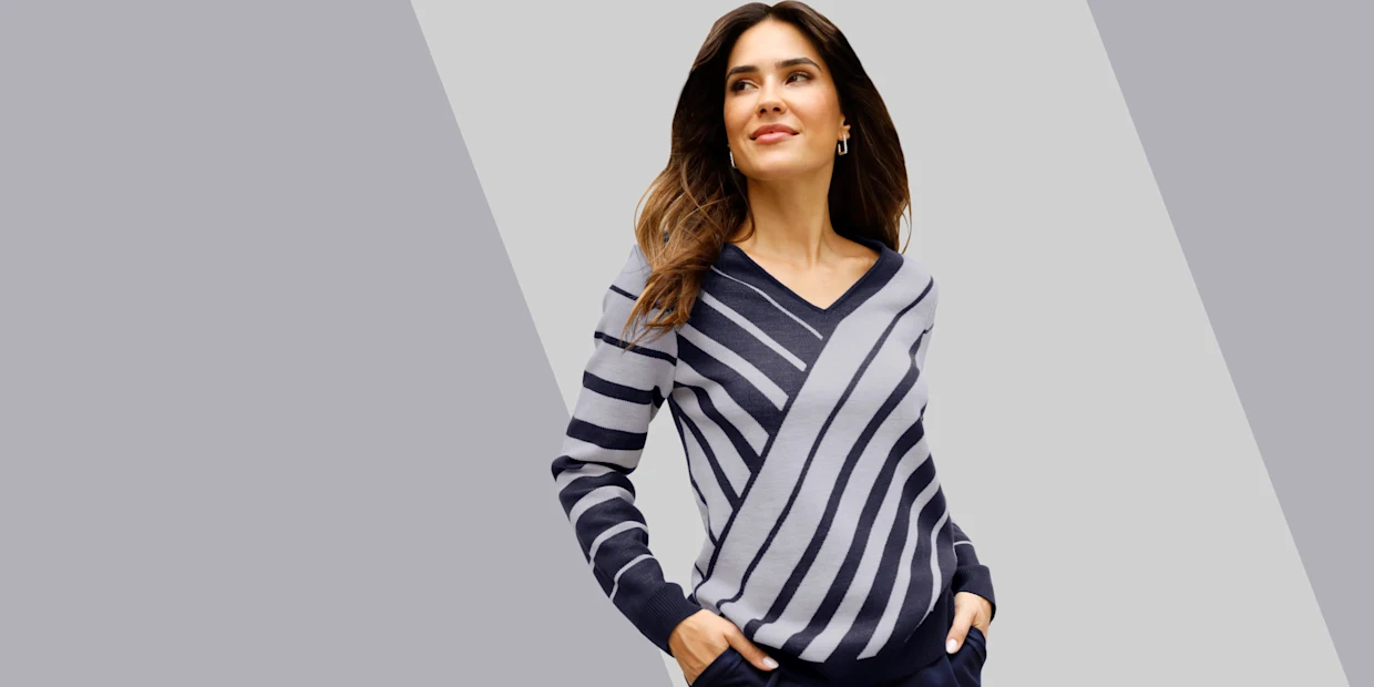 Model má na sebe sveter s výstrihom do V a diagonálnymi pruhmi v sivých a modrých odtieňoch a tmavomodré nohavice.