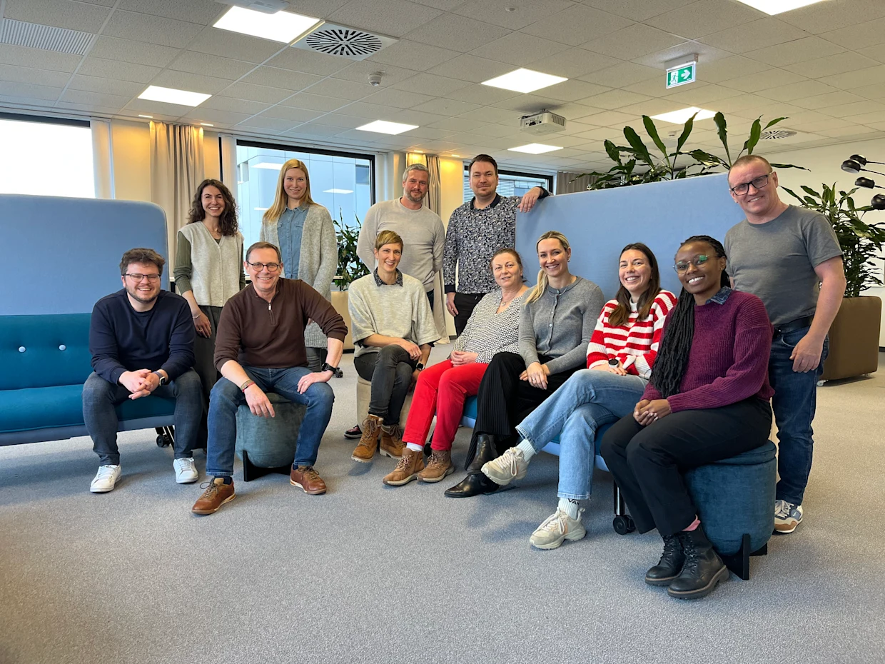 Groep mensen poseren samen in een moderne kantoorruimte met blauwe banken en groene planten op de achtergrond. Groep mensen poseren samen in een moderne kantoorruimte met blauwe banken en groene planten op de achtergrond.