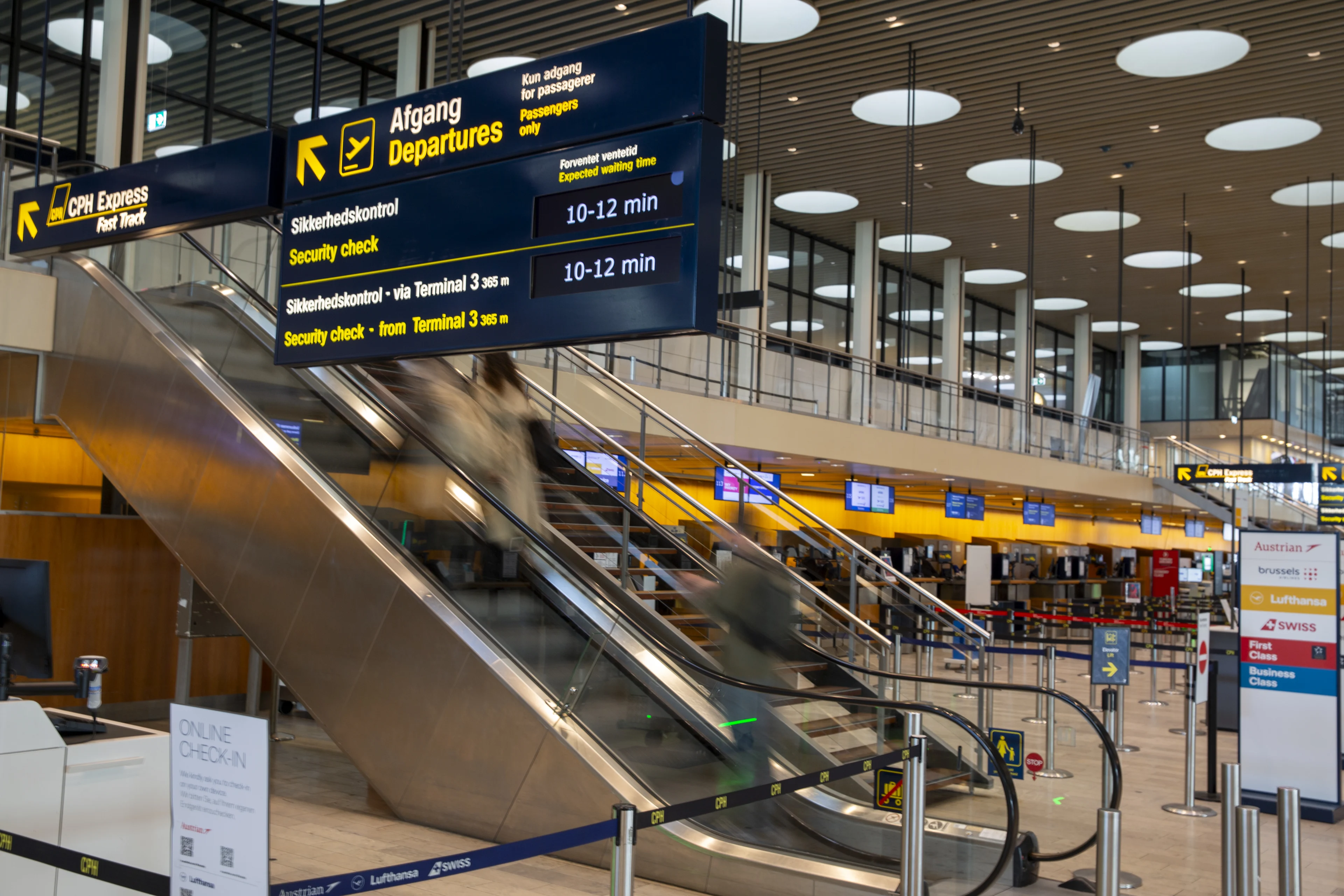 Bild från Köpenhamns Flygplats avgångshall. Man ser två rulltrappor som går upp till säkerhetskontrollen samt mörkblå informationsskyltar med gul text och flertalet incheckningsdiskar i bakgrunden.