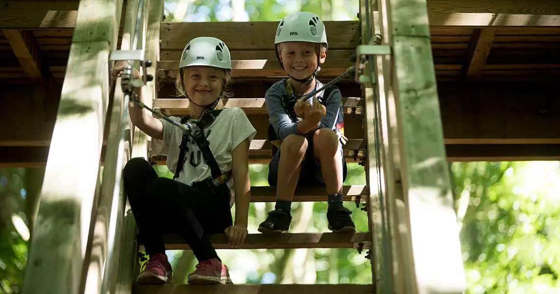 Två barn med hjälmar och säkerhetsutrustning sitter leende på en trätrappa i en äventyrsbana utomhus, omgivna av grönska.