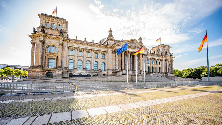 Riksdagshuset i Berlin på en solig dag. Framför byggnaden syns EU-flaggan och Tysklands flagga, lummiga gröna träd och en trottoar.
