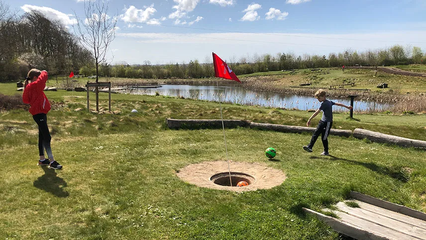 Två besökare spelar fotbollsgolf en solig dag. En pojke sparkar bollen mot ett hål som är markerat av en röd flagga. En vuxen person står bredvid och tittar på. I bakgrunden syns lite skog och en liten sjö.