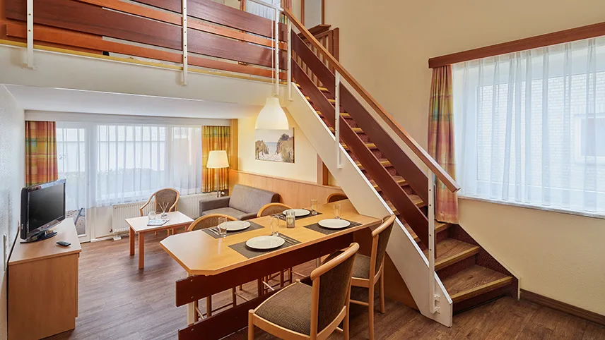 Ett av semesterhusen sett från allrummet. Man ser ett dukat bord, en soffgrupp och en trappa upp till övervåningen.