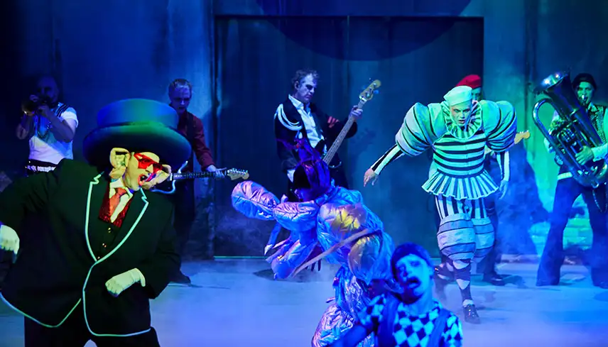 Bild på scenen med blått ljus och ett flertal skådespelare med dramatisk sminkning och färgstarka scenkonstymer som dansadr och sjunger.