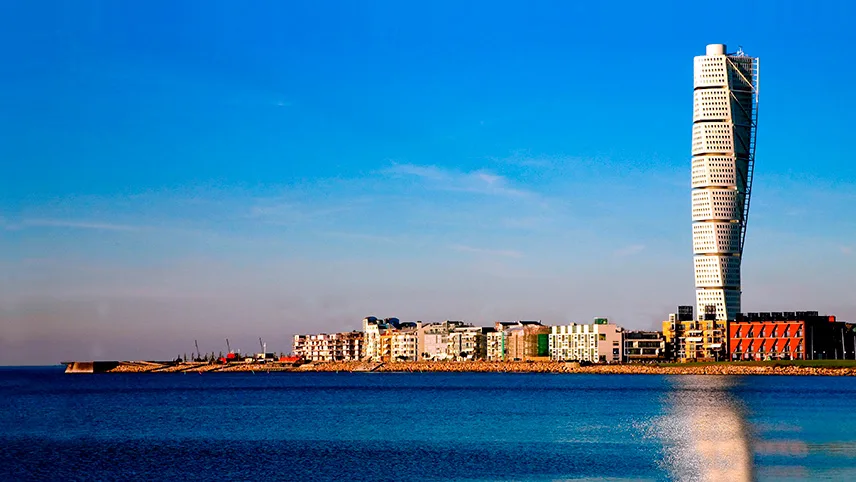 Turning Torso i Malmø tårner sig op over byen foran vandet med den blå himmel i baggrunden.