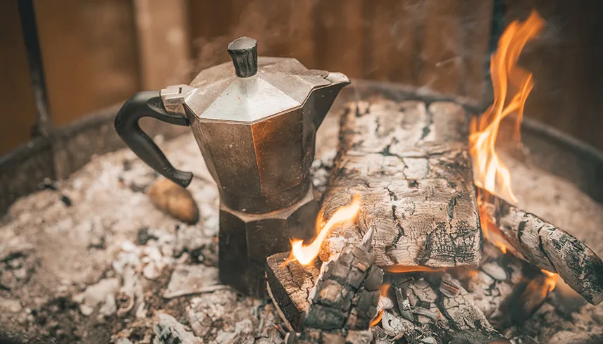 Stämningsbild med ett eldfat med björkved, eld och en Bialetti-kanna,