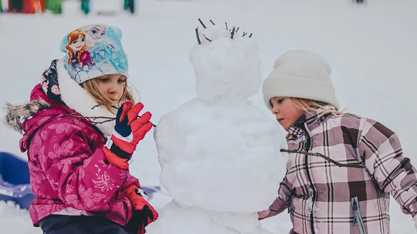 Två barn leker i snön och bygger en snögubbe med pinnar som armar och hår.