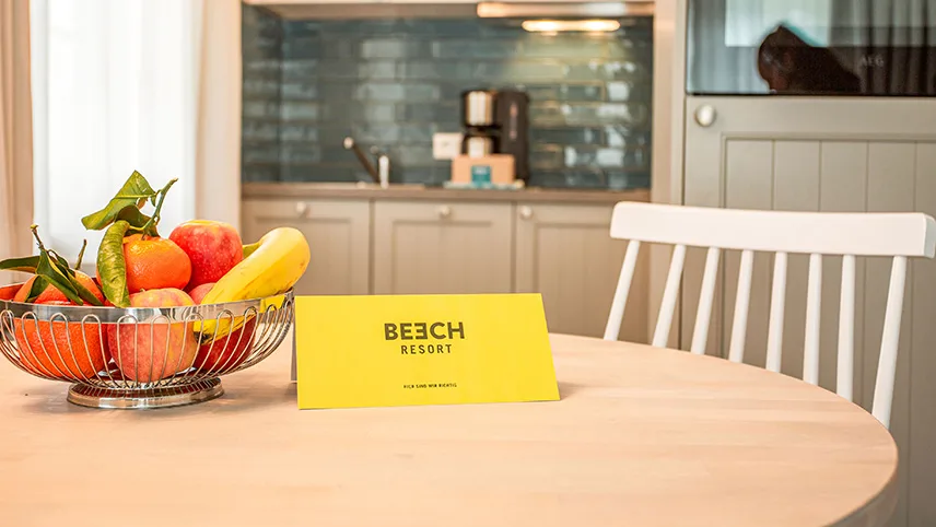 Ett kök i en av lägenheterna. I bakgrunden syns en diskbänk med kaffebryggare och i förgrunden står ett matbord med en fruktskål och ett välkomstkort med hotellets logga.
