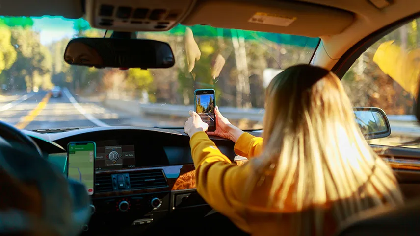 En kvinna sitter i en bil och tittar ut genom vindrutan och tar bild av vägen med sin smartphone.