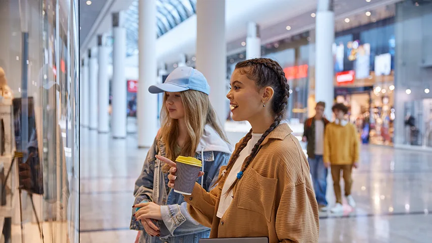 Två yngre kvinnor står framför ett skyltfönster i ett shoppingcenter och tittar och pratar.