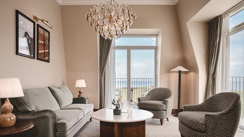 Vardagsrum från en av hotellets sviter. Man ser en soffa i dämpat grön sammet, två fåtöljer samt ett runt soffbord. I taket hänger en stor lampa och på väggarna hänger bilder. I bakgrunden syns fönster och en beige vägg.