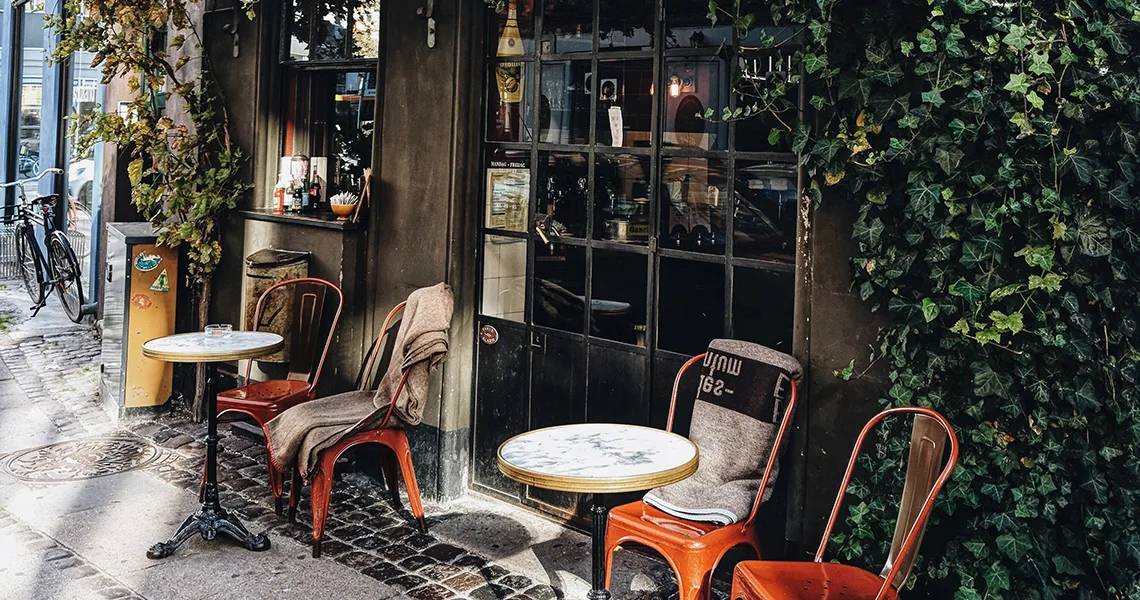 Flera stolar och små marmorbord står utanför ett café omslingrat av gröna växter längs en stadsgata med cyklar i bakgrunden.