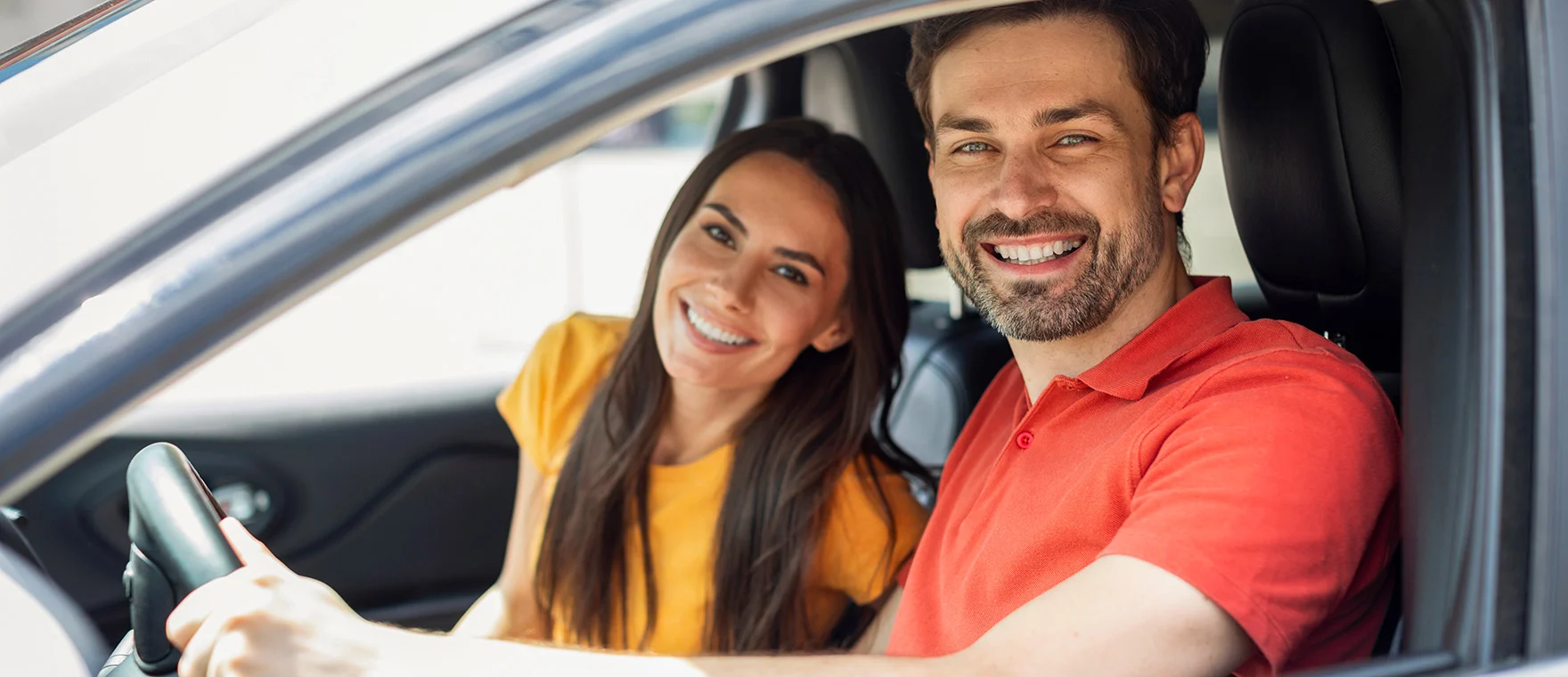 Mörkhårig man och kvinna sitter i framsätet på en grå bil och ler mot kameran.