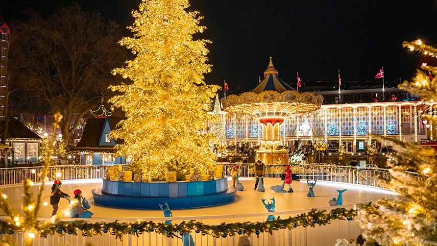 Den upplysta skridskobanan med en stor guldklädd julgran i mitten och ett flertal besökare som skridskor.