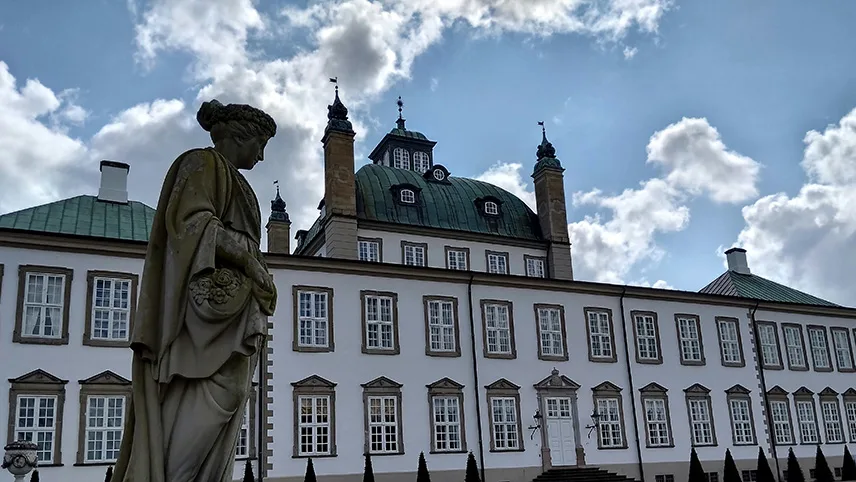 Bild av slottets fasad mot en ljusblå sommarhimmel med mjuka vita moln. I förgrunden syns en staty av en kvinna.