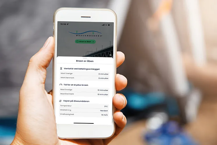 Person håller i Øresundsbrons app på mobilen och ser aktuella väntetider, körtid och väder på bron – visar hur appen ger realtidsinformation inför resan över Øresundsbron.