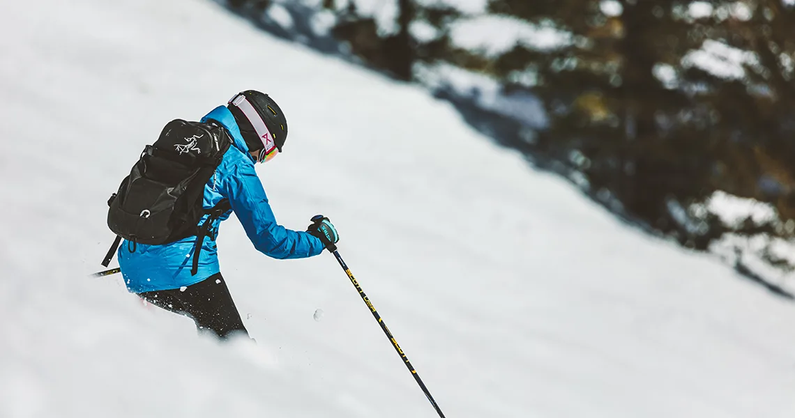 En skiløber iført blå jakke og hjelm kører ned ad en sneklædt piste i Sverige, omgivet af træer, mens de navigerer i det alpine terræn.