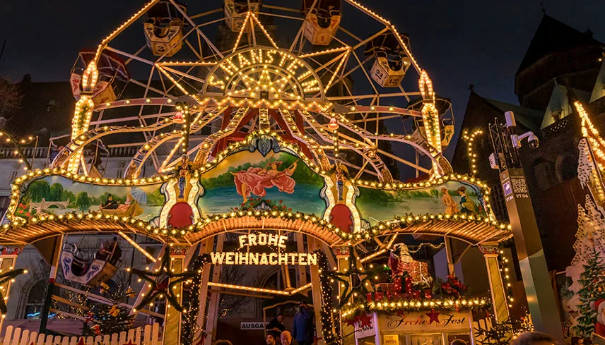 Ingången till en julmarknad i Tyskland, upplyst av varmvitt ljus med pynt och orden Frohe Weihnachten över porten.