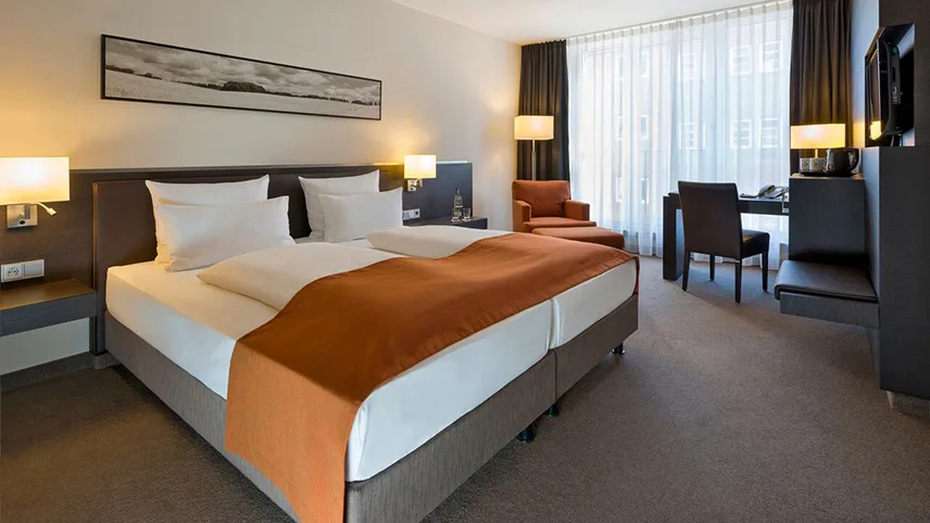 Ett av hotellets Superior-rum med dubbelsäng bäddad med vita lakan och en röd-orange pläd. Över sängen sitter en tavla och i bakgrunden ses ett skrivbord och tända lampor.