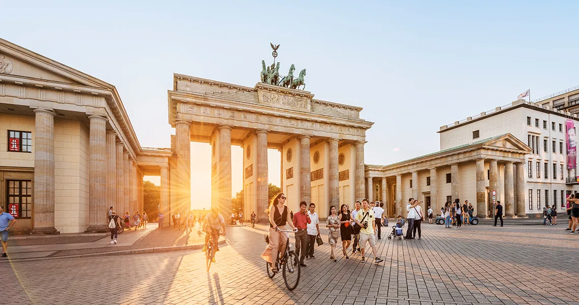 Brandenburger Tor står i solnedgången medan människor promenerar och cyklar förbi på en livlig gata i Berlin.