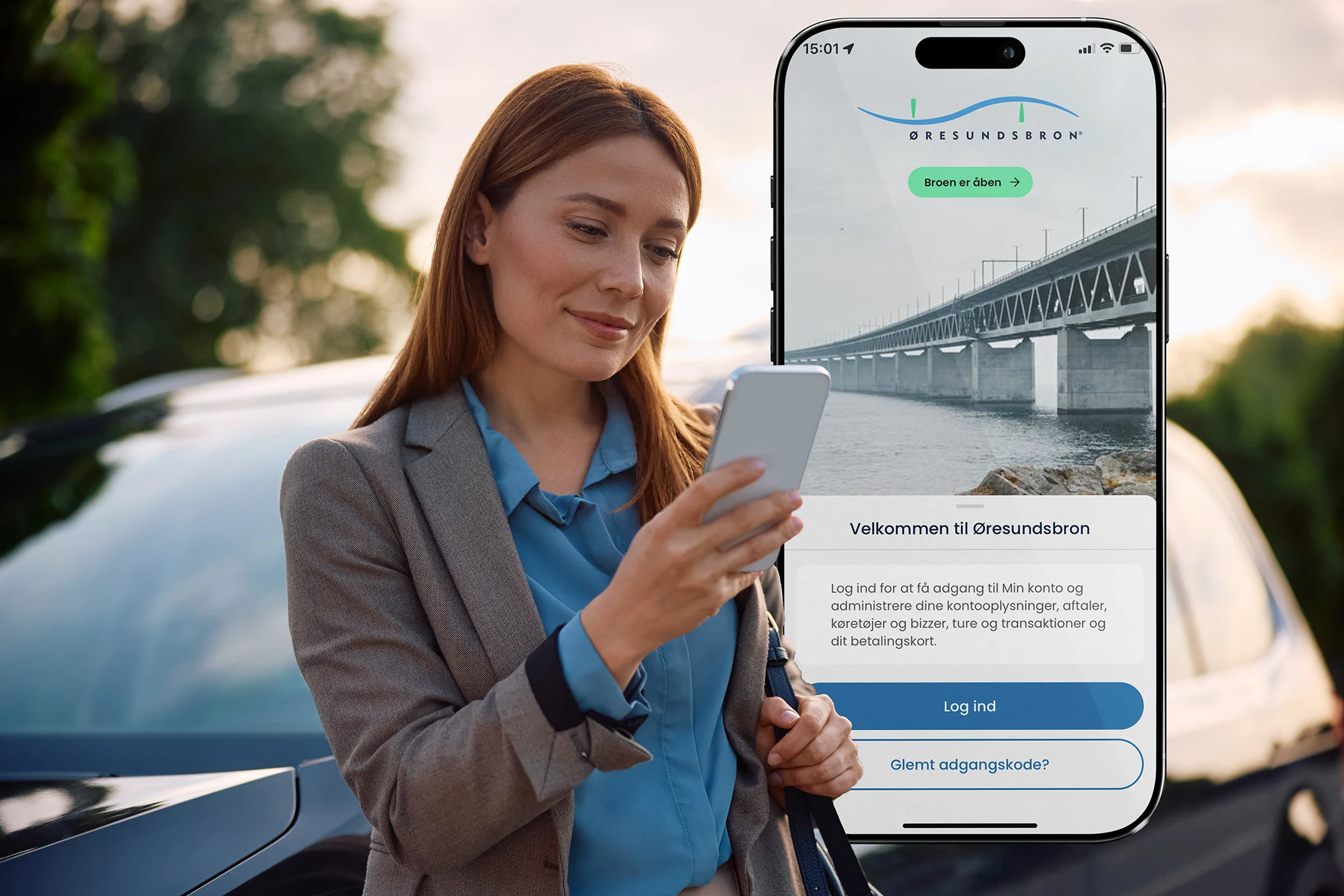 Kvinna med gul tröja och grå kappa använder Øresundsbrons app på mobilen för att planera resan och hantera sitt avtal, med bilen i bakgrunden som visar att appen förenklar allt inför körningen över Øresundsbron.