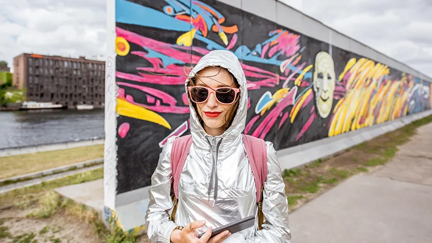 En kvinnlig resenär framför den del av Berlinmuren som har blivit ett konstmuseum och kallas East Side Gallery. I bakgrunden syns floden Spree samt muren med målningar i svart, rosa, blått och gult.
